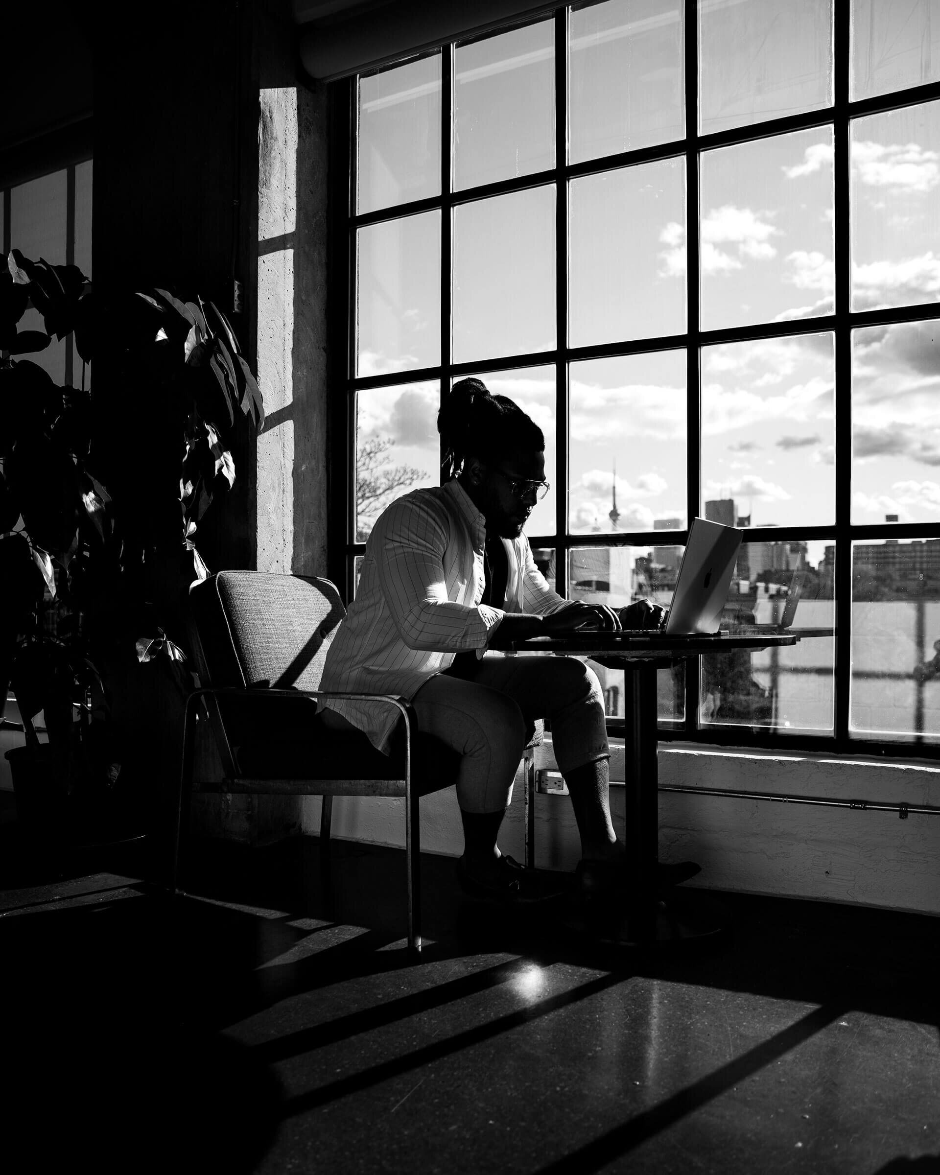 Corey working on a laptop by a tall window, in black and white